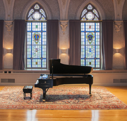 a piano in the middle of a historic concert hall space. Large stained glass windows are behind the piano.