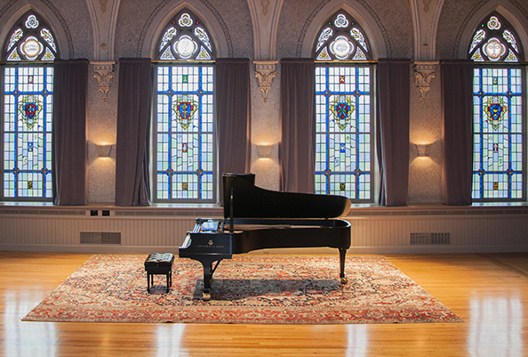 a piano in the middle of a historic concert hall space. Large stained glass windows are behind the piano.