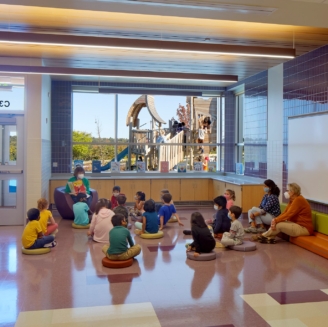 elementary school students learning in a common area of the school. in the background kids can be seen playing on a playground through the window