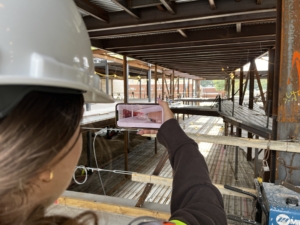 Young woman from Eureka! program looks through phone at construction site seeing rendering over actual unfinished construction