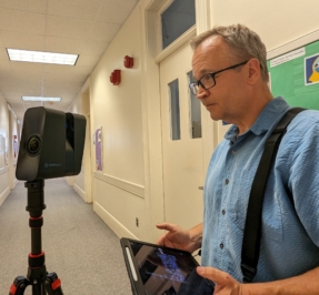 Mike stands in a hallway looking at a free standing scanner with his ipad in his hands