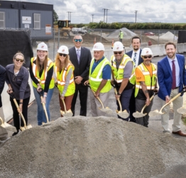 A group f people, many in hard hats and safety vests holding shovels over a large pile of dirt