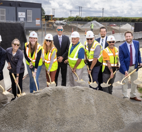 A group f people, many in hard hats and safety vests holding shovels over a large pile of dirt