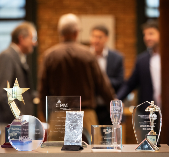 several awards in the foreground while several people are gathered in the background engaged in conversation
