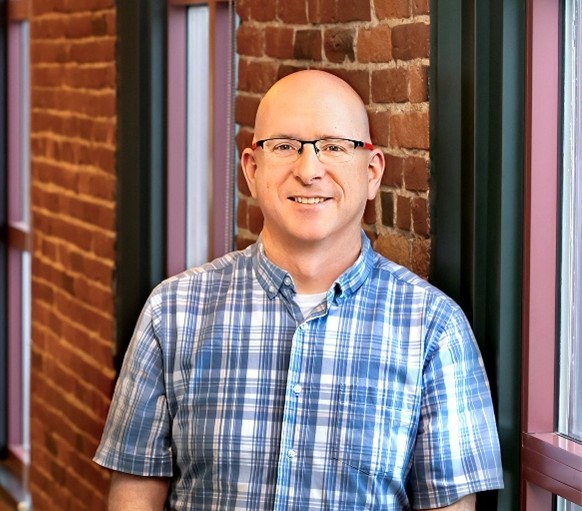 Mark Ritz stands in the office in front of the windows and brick wall
